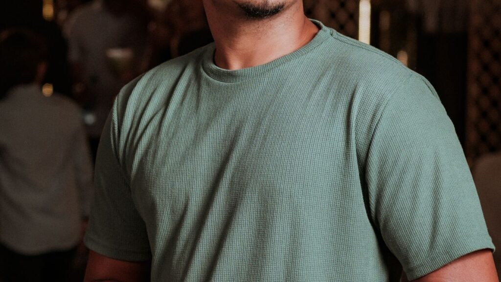 Close-up of a person wearing a sage green ribbed T-shirt at an indoor evening social event with warm ambient lighting