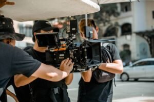 A two-person crew from a local video production company sets up a camera on a Singapore shophouse street while a colleague adjusts a reflector in the afternoon light.